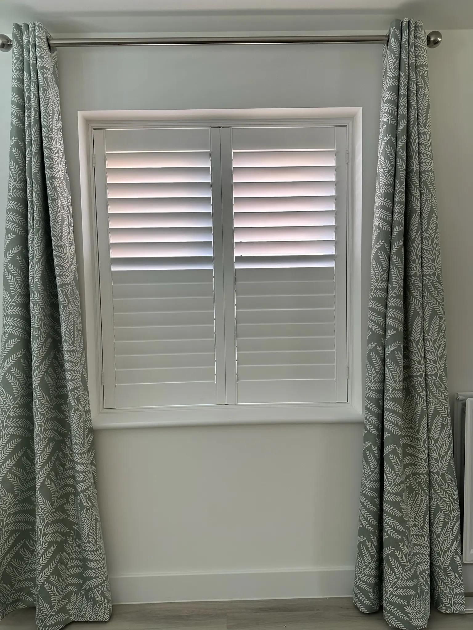 White made-to-measure shutters framed by sage green leaf-pattern curtains