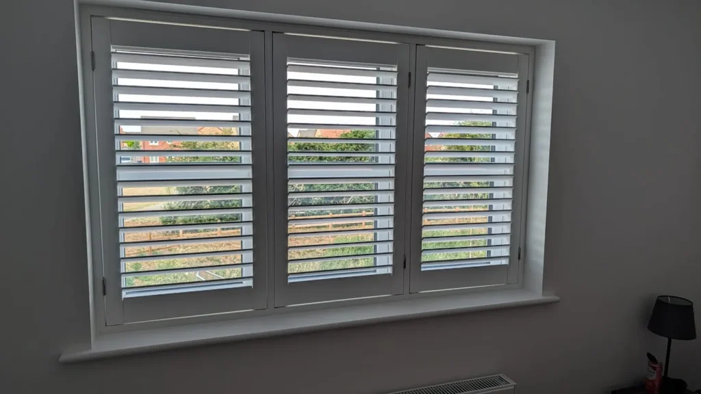 White UK-made shutters closed across a wide bedroom window, with the louvres tilted to let some daylight through